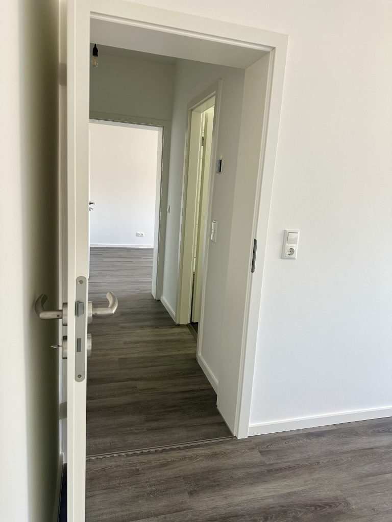 Leerer Wohnungsflur mit dunklem Laminat, durch eine Tür gesehen