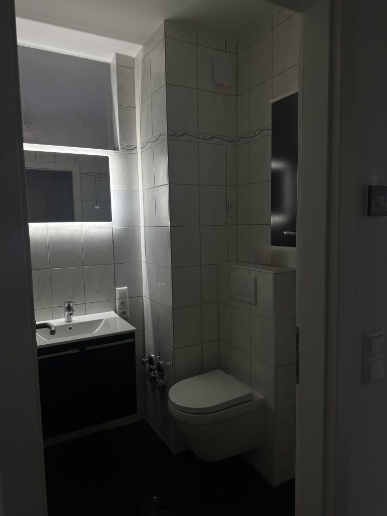 Blick aus dem Flur ins sanierte Bad mit LED-Spiegel, schwarzem Waschtisch und Wand-WC