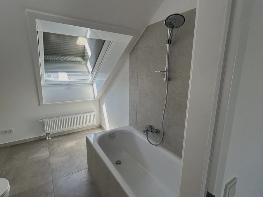 Weiße Badewanne mit Handbrause vor beigem Fliesenspiegel unter einem Dachfenster