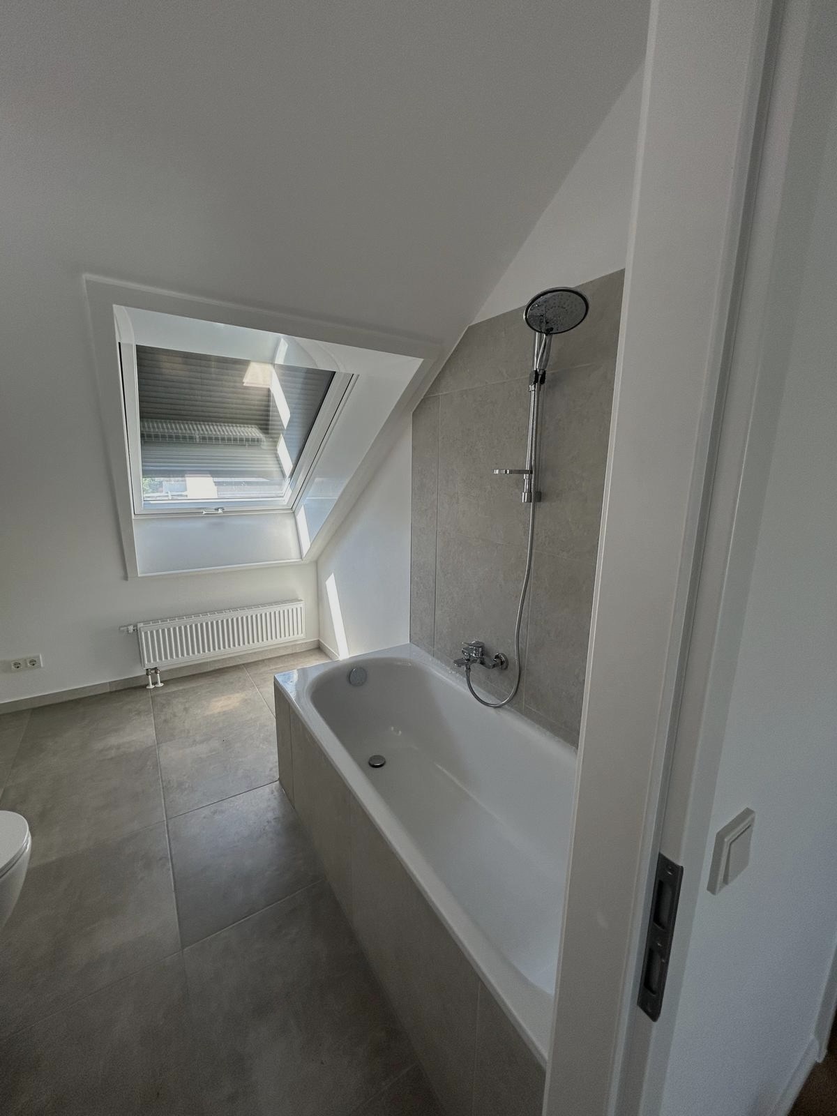 Weiße Badewanne mit Handbrause vor beigem Fliesenspiegel unter einem Dachfenster