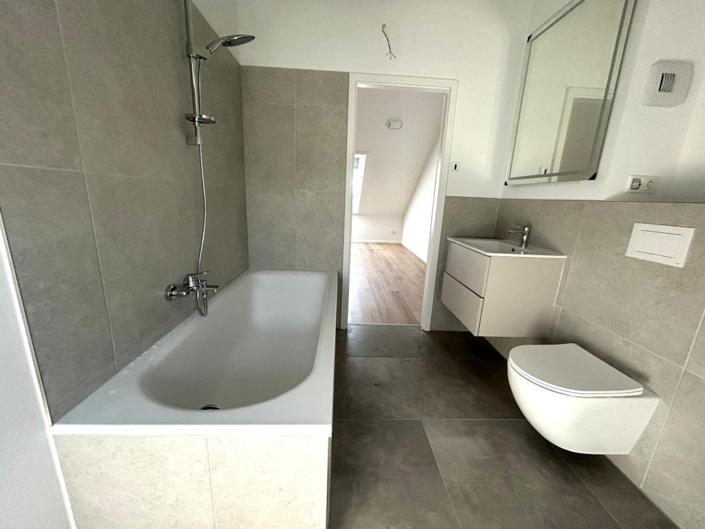 Bad mit Badewanne, Regendusche, weißem Waschtisch und Wand-WC in grauen Fliesen