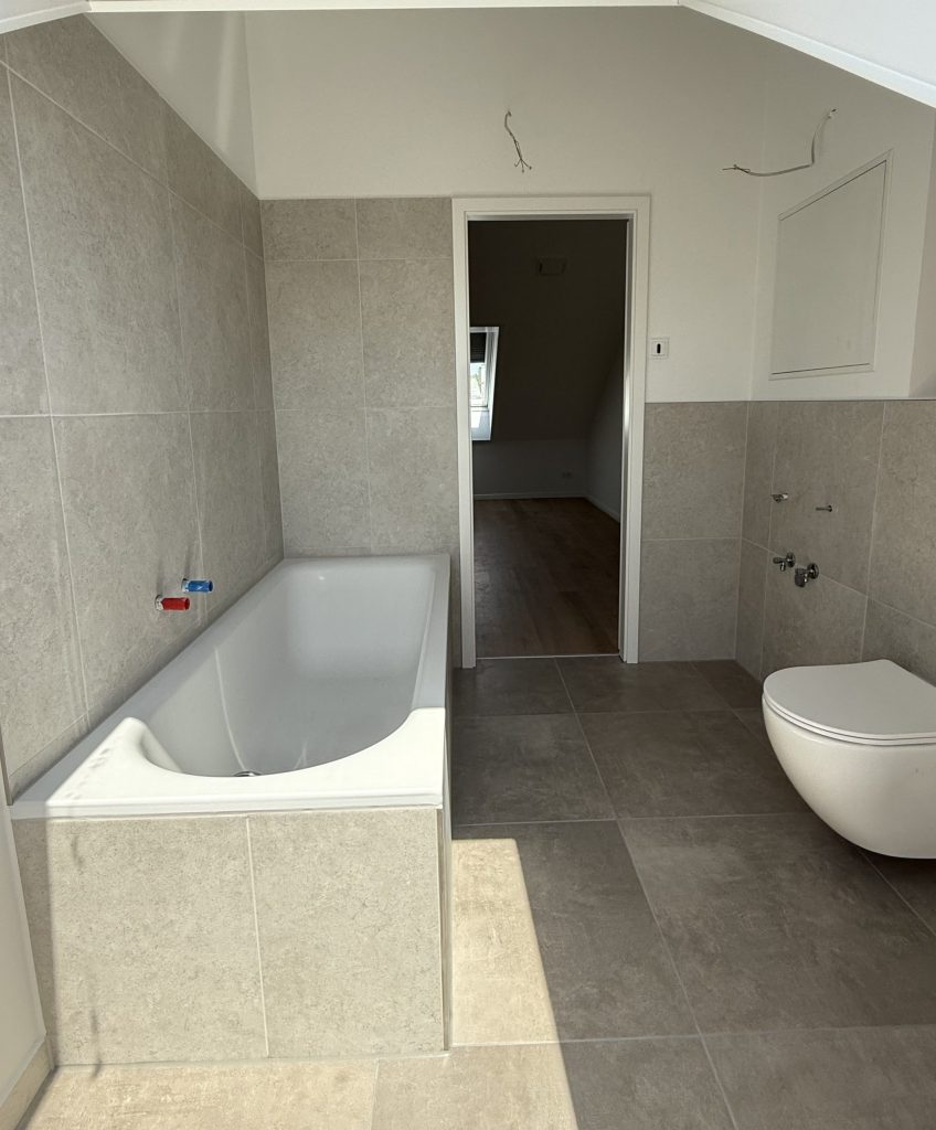 Bad mit eingebauter Badewanne, Wand-WC und offener Tür zum Nebenraum