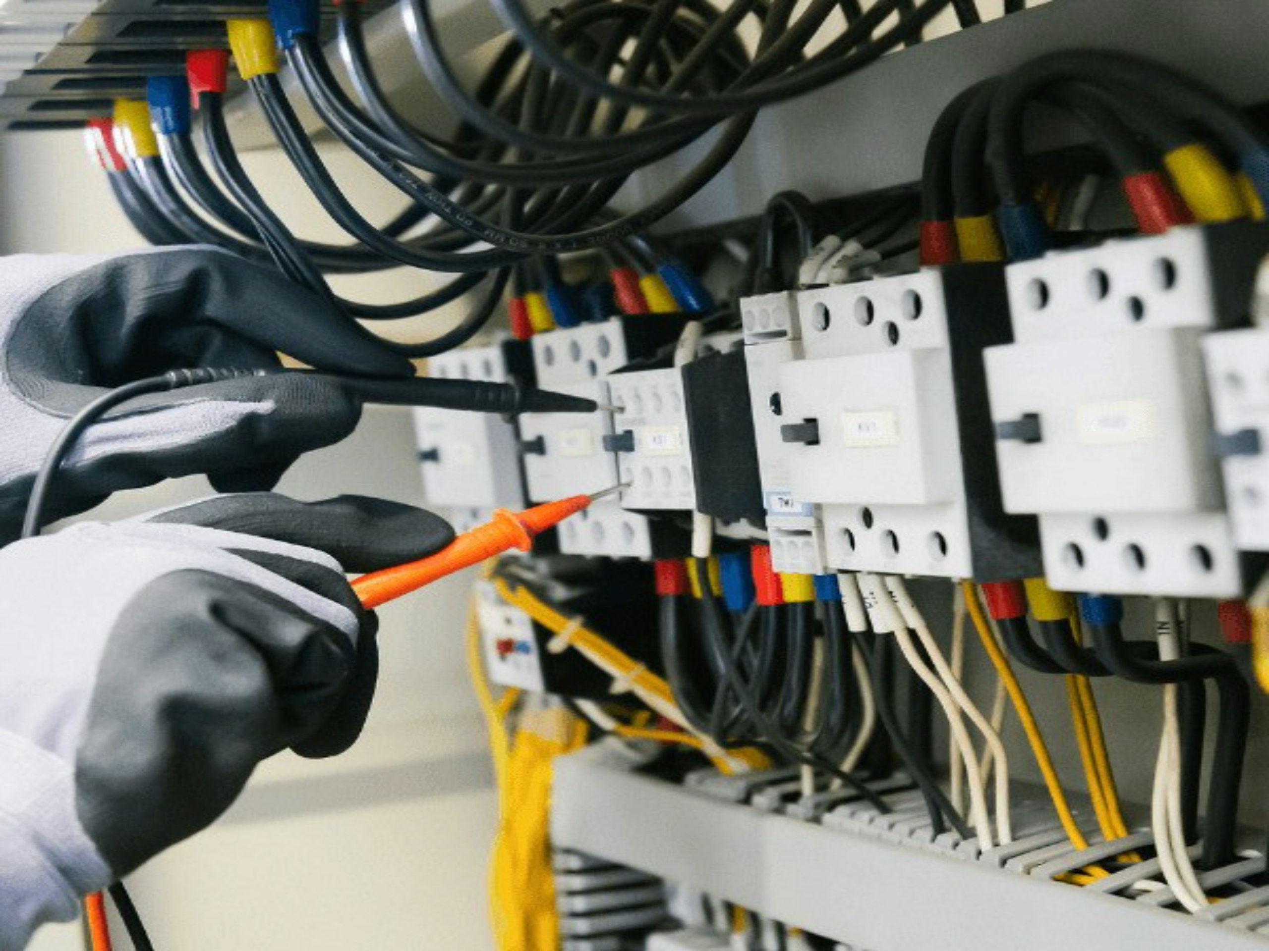 Behandschuhte Hände prüfen mit einem Messgerät die Klemmen einer Elektroverteilung