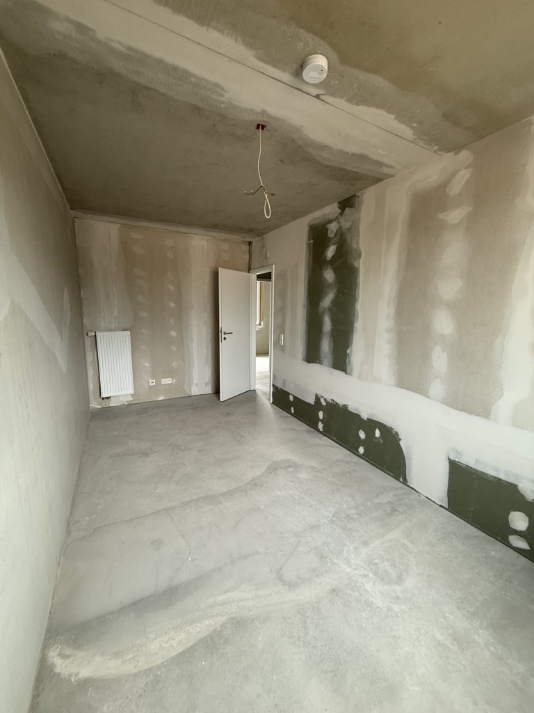 Zimmer im Rohbau mit Heizkörper, offener Tür und Feuchtraumplatten am Sockel