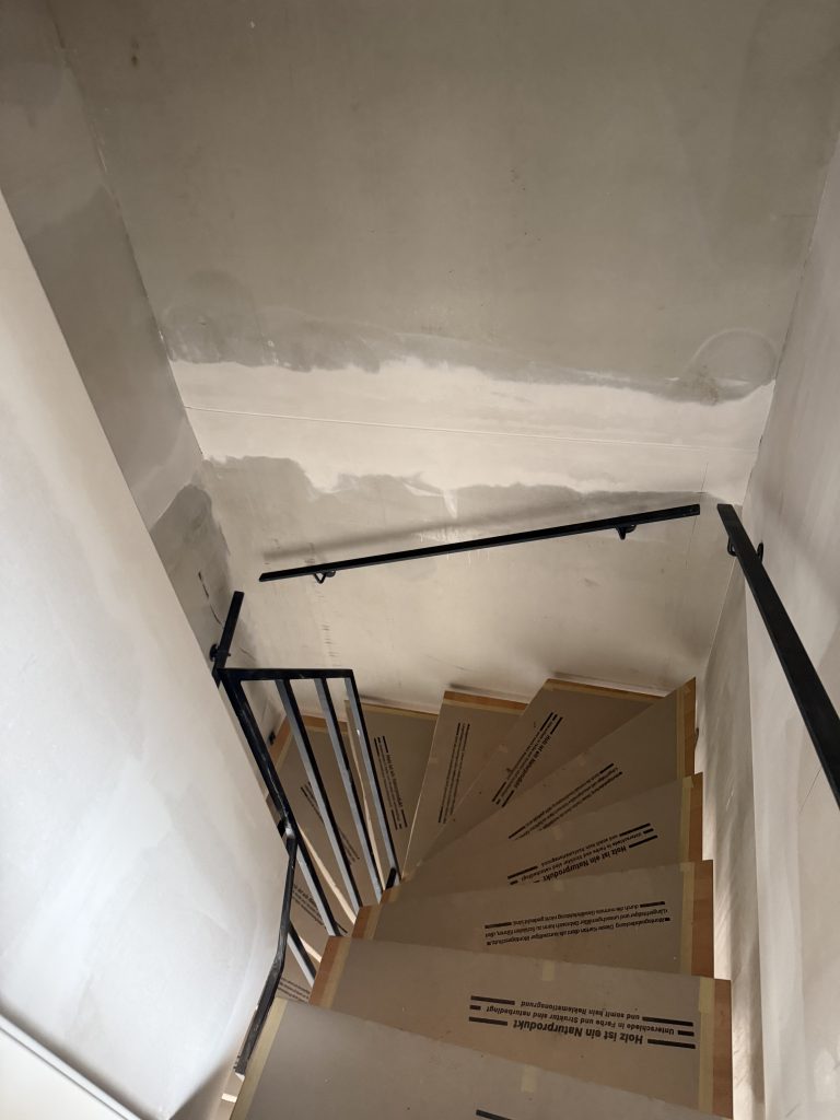 Blick von oben auf eine Treppe mit abgedeckten Holzstufen und schwarzem Geländer