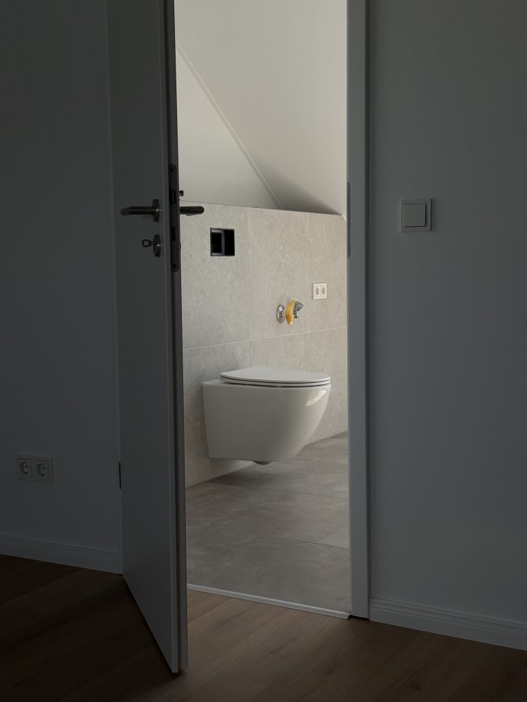 Blick durch eine angelehnte Tür auf ein Wand-WC im gefliesten Bad